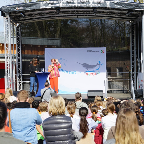 Frau mit buntem Outfit spricht auf einer Bühne vor einer Gruppe von Kindern und Erwachsenen bei einer Outdoor-Veranstaltung.