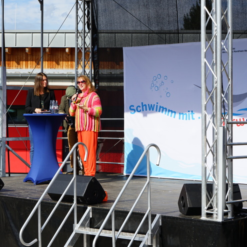 Zwei Frauen stehen auf einer Bühne, während eine der beiden ein Mikrofon hält und spricht. Im Hintergrund ein plakatives Banner.