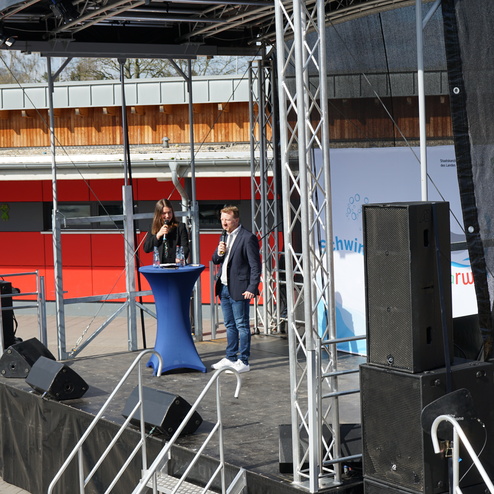 Zwei Personen stehen auf einer Bühne, während einer von ihnen ein Mikrofon hält und spricht. Im Hintergrund sind Lautsprecher zu sehen.