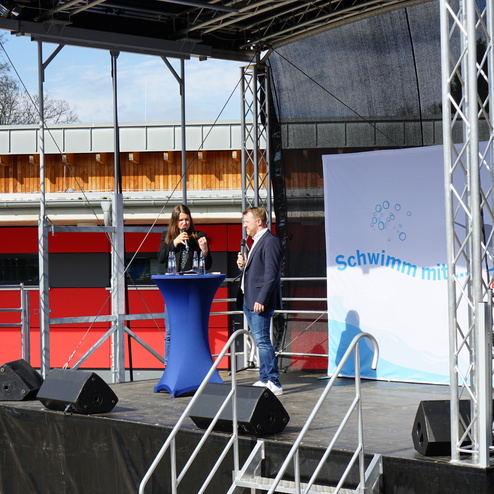 Zwei Personen stehen an einem Tisch auf einer Bühne mit Mikrofonen; im Hintergrund ein Banner mit dem Schriftzug "Schwimm mit...".