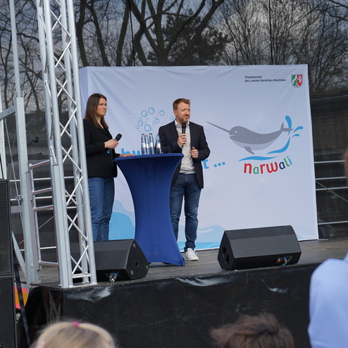 Zwei Sprecher stehen an einem Tisch auf einer Bühne vor einem Banner mit einem Narwal-Motiv.