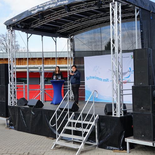 Zwei Personen stehen auf einer Bühne vor einem Banner mit dem Text „Schwimm mit...“ und Rednerpult in einer Außenanlage.