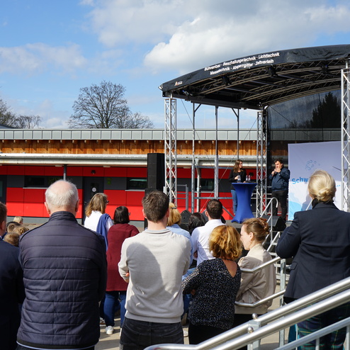 Eröffnungsrede vor Publikum auf einem Außengelände mit Bühne und rot-grauer Schulgebäude im Hintergrund.