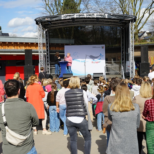 Menschenmenge schaut auf eine Bühne mit einer Präsentation im Freien, umgeben von Bäumen und Gebäuden.