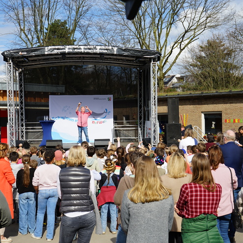 Bühne mit einem Redner und Publikum aus Kindern und Erwachsenen bei einer Veranstaltung im Freien.