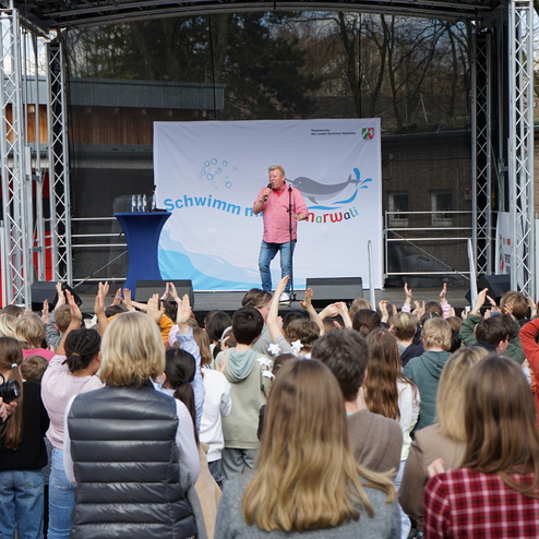 Mann mit Mikrofon steht auf einer Bühne, umgeben von einer Zuschauermenge von Kindern und Erwachsenen.