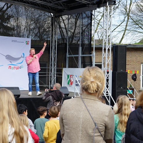 Ein Redner steht auf einer Bühne und spricht zu einer Gruppe von Kindern bei einer Veranstaltung im Freien.