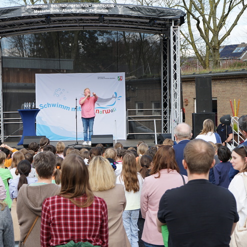 Ein Redner steht auf einer Bühne und spricht zu einer Gruppe von Kindern und Erwachsenen bei einer Veranstaltung.