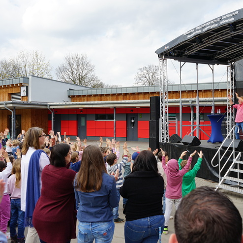Kinderschar hebt Hände zu einem Darbietung auf einer Bühne in einem Schulhof mit modernen Gebäuden im Hintergrund.
