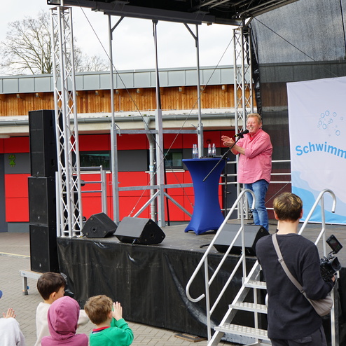Mann spricht auf einer Bühne vor einer Gruppe Kinder, während eine beflaggte Wand im Hintergrund sichtbar ist.