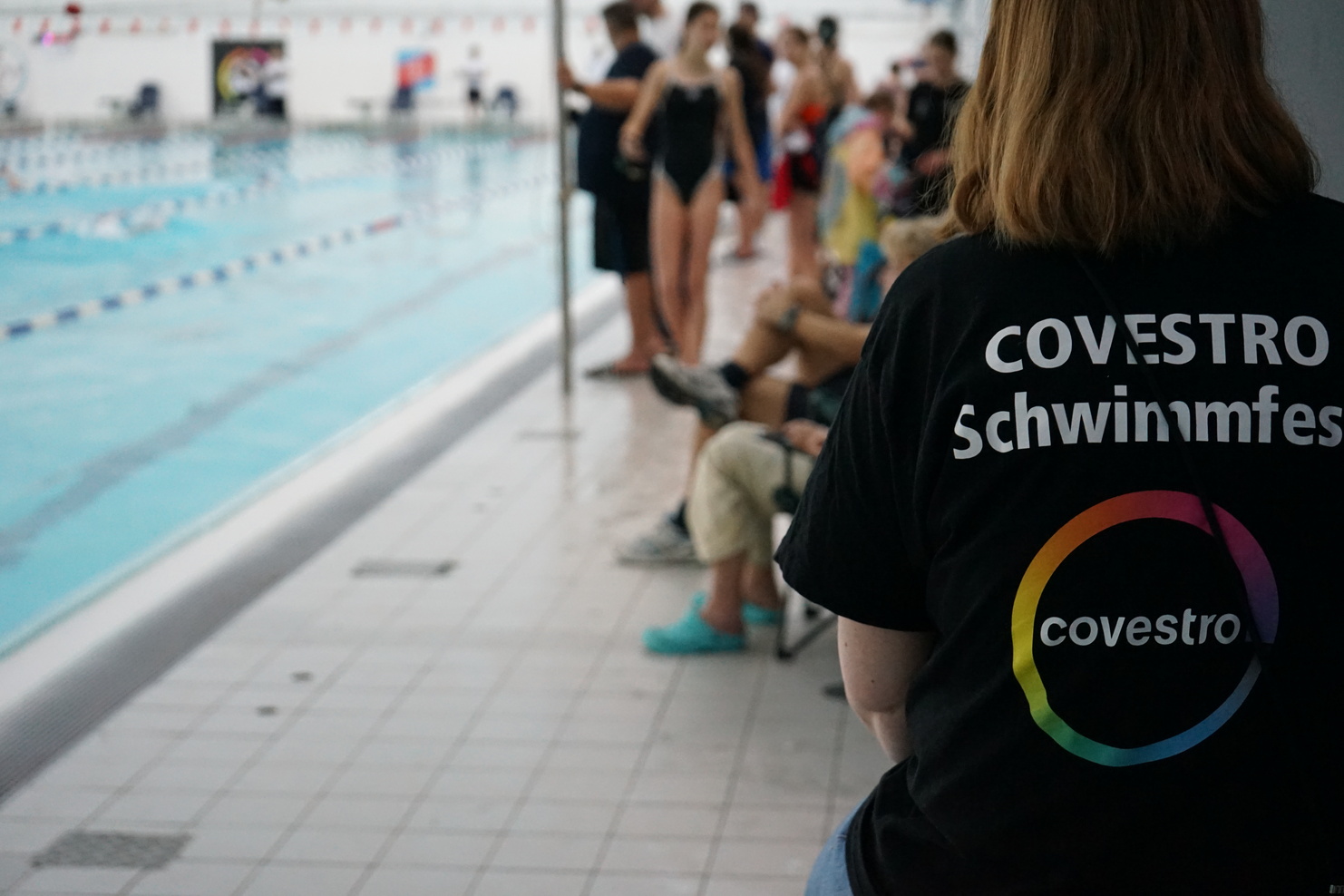 Person mit COVESTRO-Schwimmfest-T-Shirt sitzt am Beckenrand eines Schwimmbeckens mit Zuschauern im Hintergrund.
