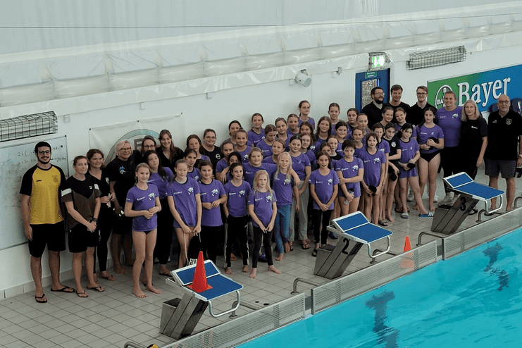 Gruppenfoto von Schwimmern und Trainern in lila Shirts am Beckenrand eines Schwimmbades mit Startblöcken.