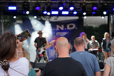 Frau filmt eine Band auf der Bühne, während Zuschauer im Vordergrund aufmerksam zuschauen. Buntes Lichtschein auf Bühne.