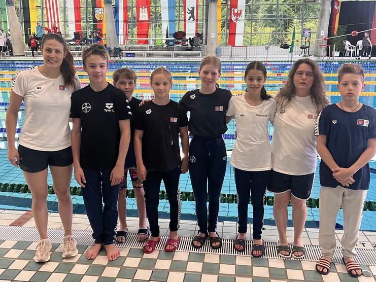 Gruppe von Schwimmern, bestehend aus einer Trainerin und sechs Kindern, vor einem Schwimmbecken mit Flaggen im Hintergrund.