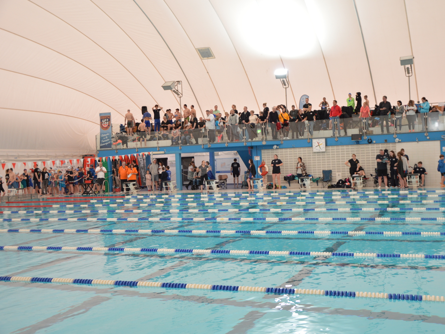Schwimmhalle mit vielen Zuschauern auf der Tribüne und mehreren Bahnmarkierungen im Wasser.