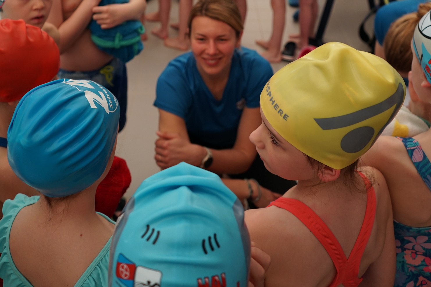 Gruppe von Kindern in Badeanzügen und Badekappen wartet an einem Schwimmbecken, während Trainerin Anweisungen gibt.