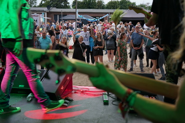 Bühnenansicht mit Bandmitgliedern im Vordergrund und Publikum, das bei einem Konzert auf einer Freiluftveranstaltung feiert.