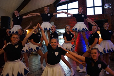 Gruppe von Tänzerinnen in blauen und weißen Kostümen, die eine Formation auf einer Bühne präsentieren.