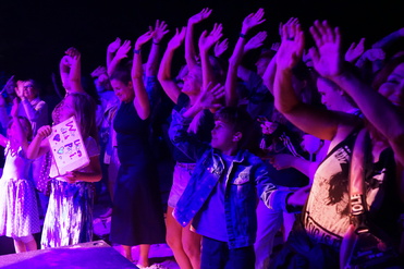 Begeisterte Menschen mit erhobenen Händen feiern auf einer Bühne, umgeben von lila Licht. Ein Kind hält ein Schild hoch.