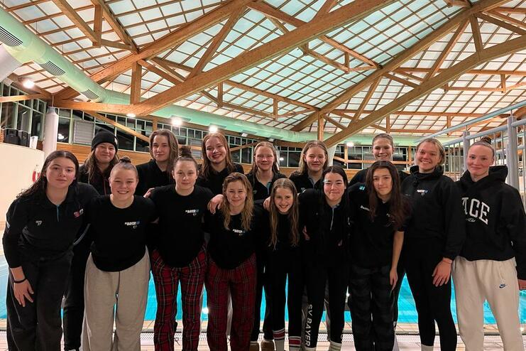 Gruppe von 15 weiblichen Schwimmerinnen posiert am Beckenrand in einem Hallenbad mit Holzstruktur.