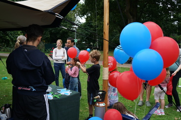 Kinder spielen auf einer Wiese mit roten und blauen Luftballons, während Erwachsene in der Nähe stehen und zuschauen.