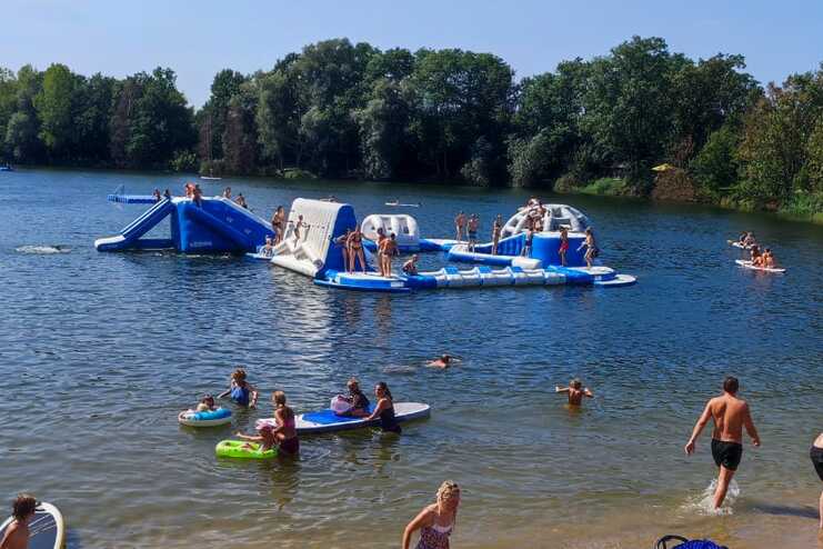 Menschen am Strand spielen im Wasser und auf Sand, während im Hintergrund Wasserspielgeräte zu sehen sind.