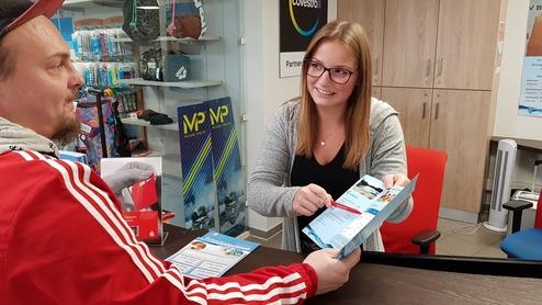 Frau an einem Empfangstresen erklärt einem Mann Informationen aus einem Flyer in einem Verkaufsraum.