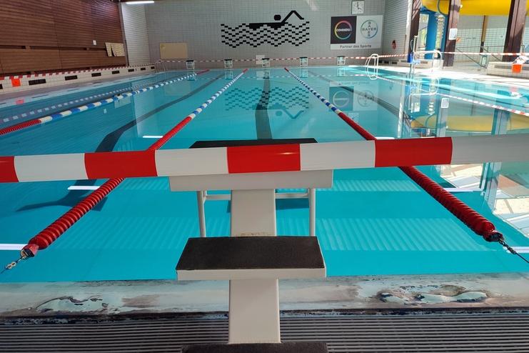 Blick auf ein Schwimmbecken mit Startblock und roten Absperrb&auml;ndern, umgeben von blauen Wasserbahnen.