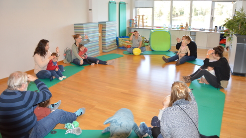 Gruppe von Erwachsenen mit Kindern in einem Raum, die auf grünen Matten sitzen und sich aktiv unterhalten.