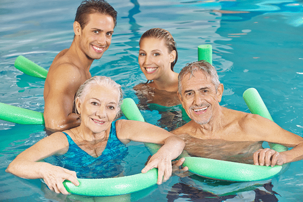 Vier Personen im Wasser halten Schwimmnudeln, lächeln und posieren gemeinsam in einem Schwimmbad.