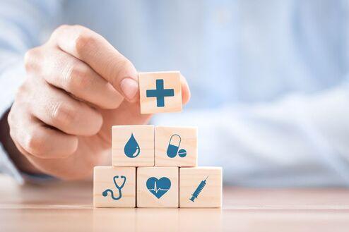 Hand, die einen Holzblock mit einem Plus-Symbol hält; im Hintergrund gestapelte Blöcke mit medizinischen Symbolen.