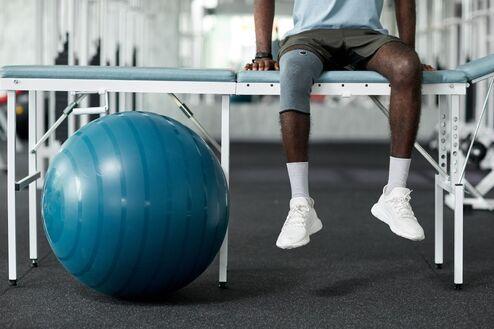 Person sitzt auf einer Bank im Fitnessstudio, daneben ein blauer Gymnastikball auf dem Boden.