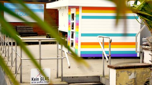 Bunter Beach Lounge mit Sitzbank, Sichtschutz und einem Hinweis auf ein Glasverbot in der Nähe einer Palmenspalte.