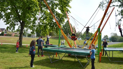Kinder springen auf Trampolinen unter einem großen Trampolin-Gerüst in einem Park mit Bäumen im Hintergrund.
