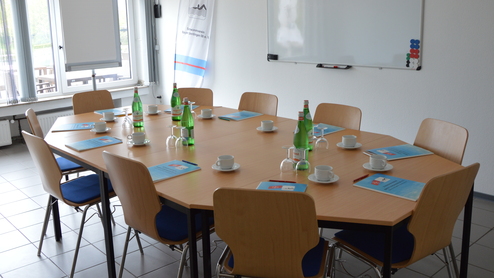 Konferenzraum mit ovalem Tisch, Wassergläsern, Kaffeetassen und Informationsmaterialien, beleuchtet von natürlichem Licht.