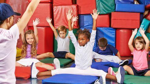 Kids im Sportraum sitzen auf Matten, heben die H&auml;nde und folgen einer Anleitung von einem Trainer.