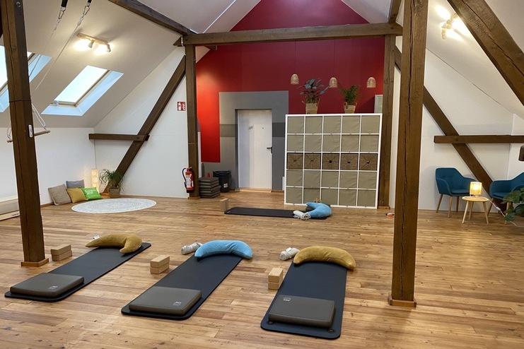 Geräumiger Yoga-Raum mit Matten, Kissen und Holzfußboden, kombiniert mit gemütlicher Sitzecke und Pflanzen.