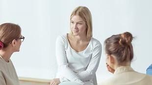 Vier Personen sitzen in einem hellen Raum im Gespräch, eine Frau in der Mitte hört aufmerksam zu.