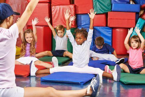 Kids im Sportraum sitzen auf Matten, heben die Hände und folgen einer Anleitung von einem Trainer.