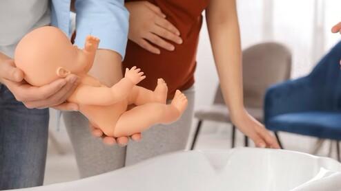 Person hält eine Plastikbabypuppe über eine weiße Badewanne, während zwei weitere Personen daneben stehen.