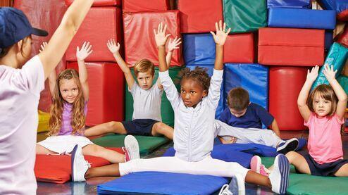 Kids im Sportraum sitzen auf Matten, heben die Hände und folgen einer Anleitung von einem Trainer.