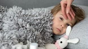 Krankes Kind mit einem Schmusetier liegt unter einer grauen Decke, während eine Hand seine Stirn berührt.