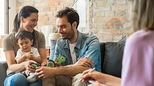 Familie sitzt auf einem Sofa, während ein Mann mit einem Kind spielt und eine Frau Notizen macht. Hintergrund mit Ziegelmauer.