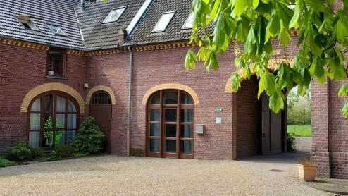 Zwei Backsteinbauten mit großen Fenstern und einem Tor im Innenhof, umgeben von frischem Grün und Kiesweg.