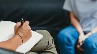 Nahaufnahme einer Hand, die in ein Notizbuch schreibt, während eine Person auf einem Sofa sitzt.