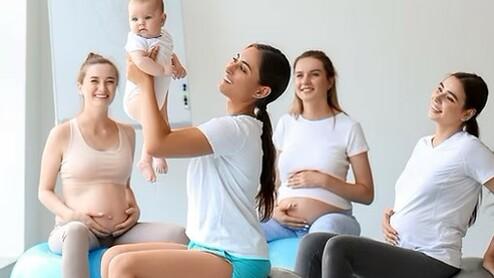 Vier schwangere Frauen sitzen auf Gymnastikbällen, während eine Frau ein Baby lächeln hält. Helle, freundliche Umgebung.