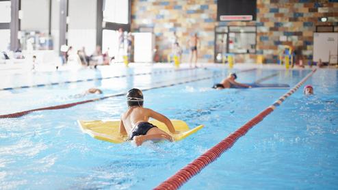 Ein Kind schwimmt mit einem gelben Schwimmbrett in einem Hallenbad, während andere Schwimmer im Hintergrund trainieren.