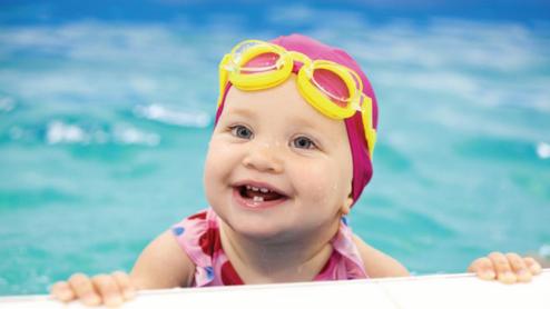 Lächelndes Kleinkind mit gelben Schwimmbrillen und rotem Badeanzug am Rand eines Schwimmbeckens.