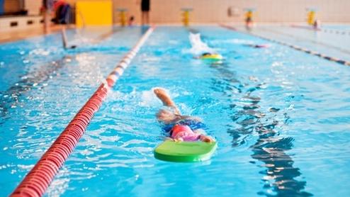 Junge schwimmt mit einem Schwimmbrett in einem Hallenbad, umgeben von blauen Wasser und roten Bahnmarkierungen.