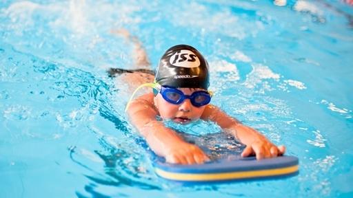 Ein Kind schwimmt mit einer Schwimmbrette in einem blauen Schwimmbecken, tr&auml;gt eine Badekappe und Schwimmbrille.
